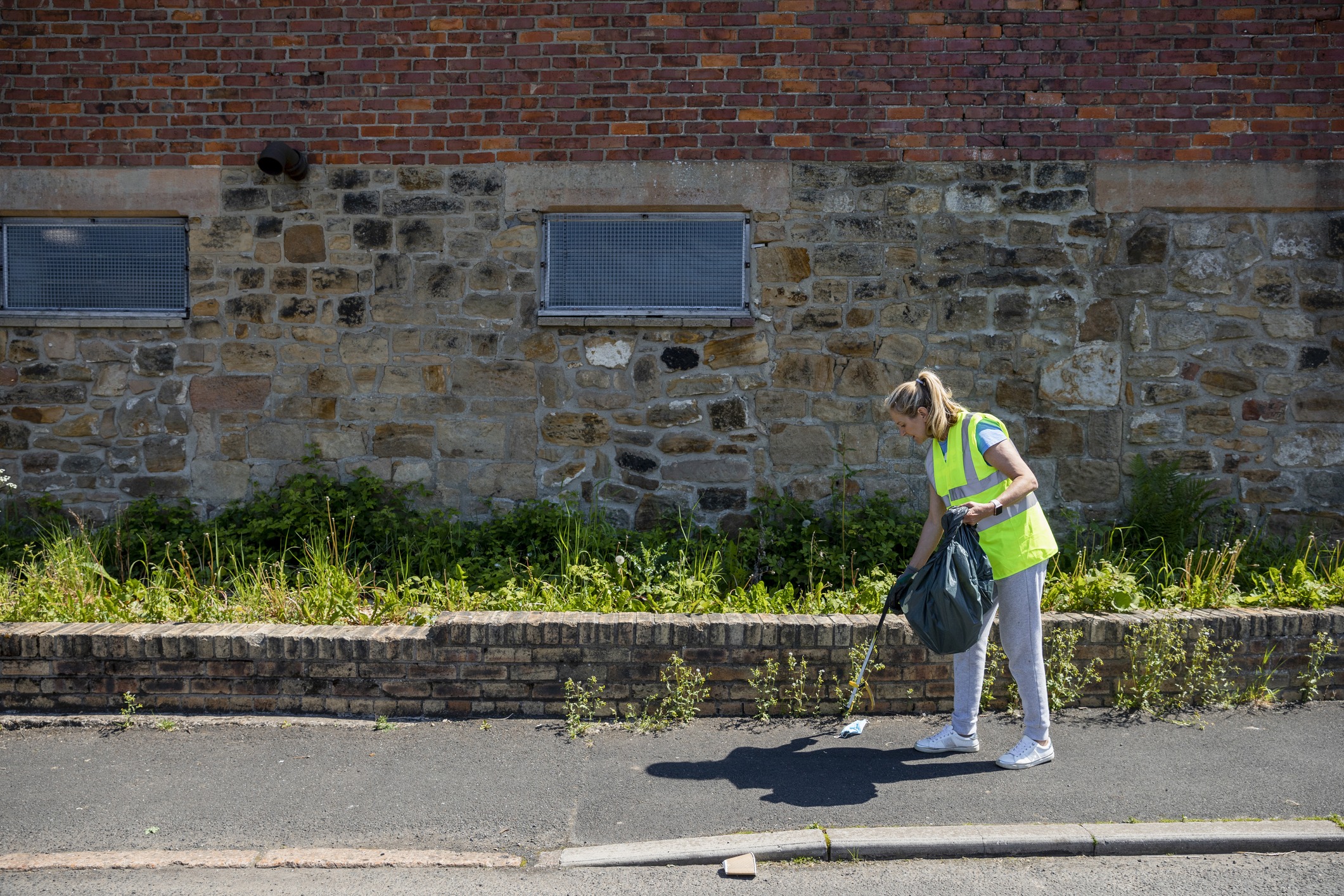 Side view of a woman litter picking in her community in the North East of England. She is wearing a high vis jacket and is using a grabber tool to put the litter into a garbage bag.