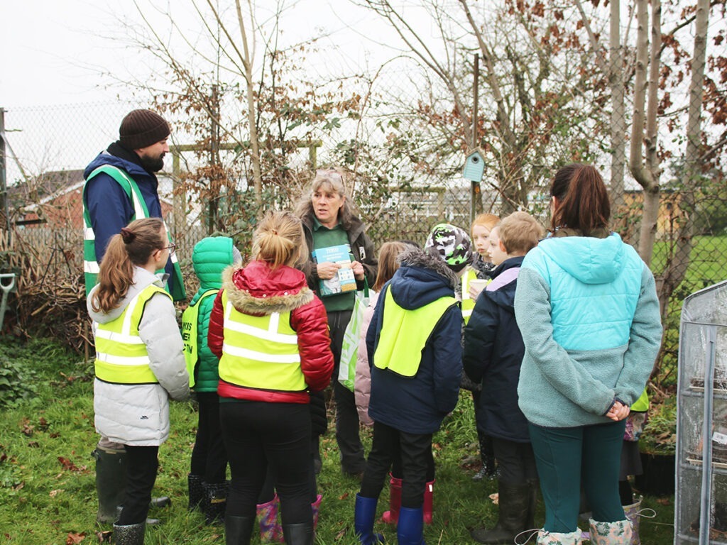 School children gathered round teacher in community garden listening to teacher talk about wildlife