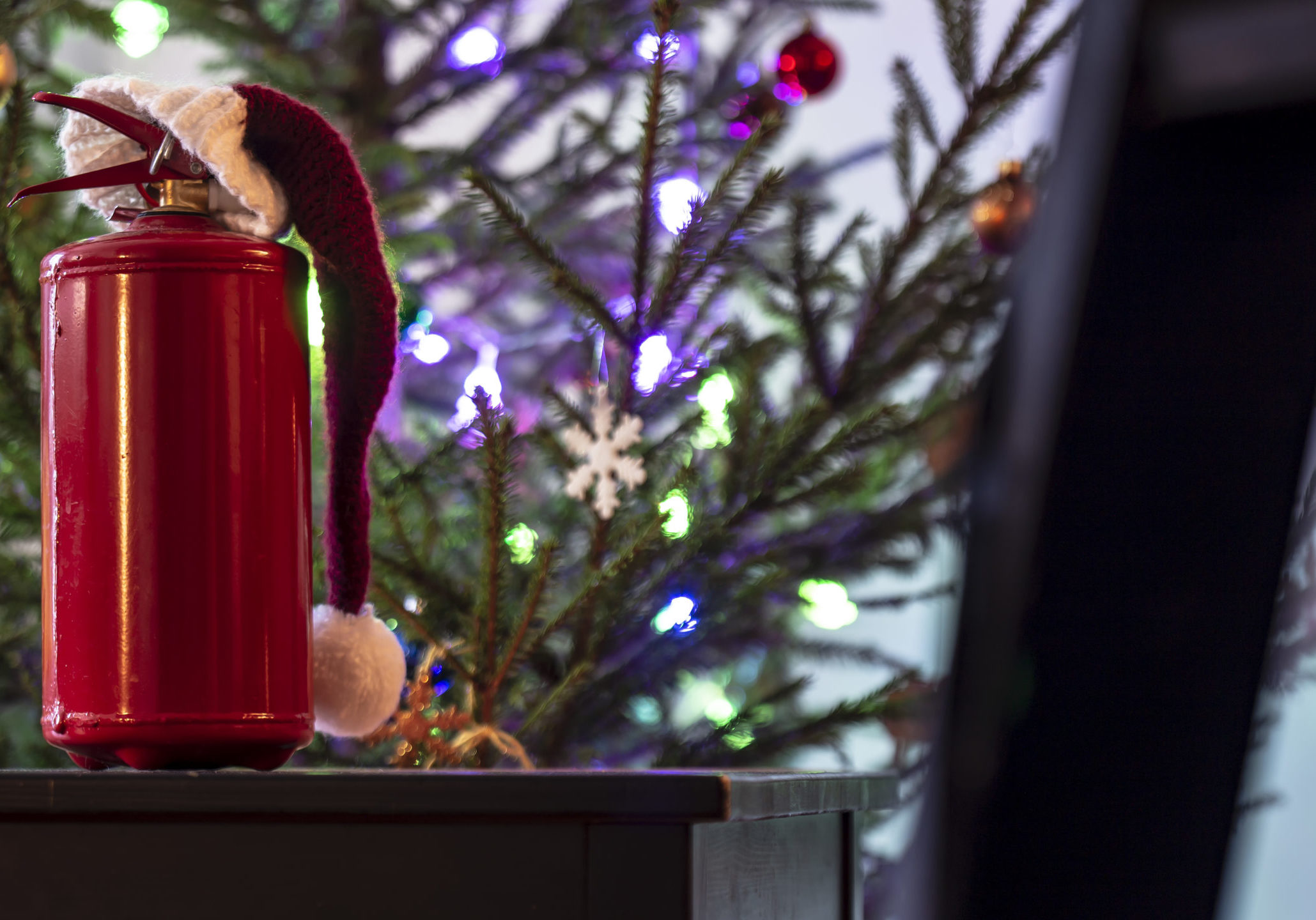 Extinguisher in red cap of Santa Claus standing on a black wooden table against the background of the Christmas tree, decorated with colorful garlands and toys, in a homelike atmosphere. Safety concept.